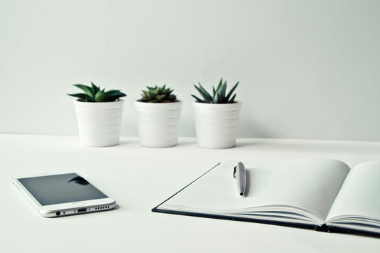 A white desk with a note book open at a blank page, with a pen sat on it. There is also a mobile phone and three small plants on the desk.
