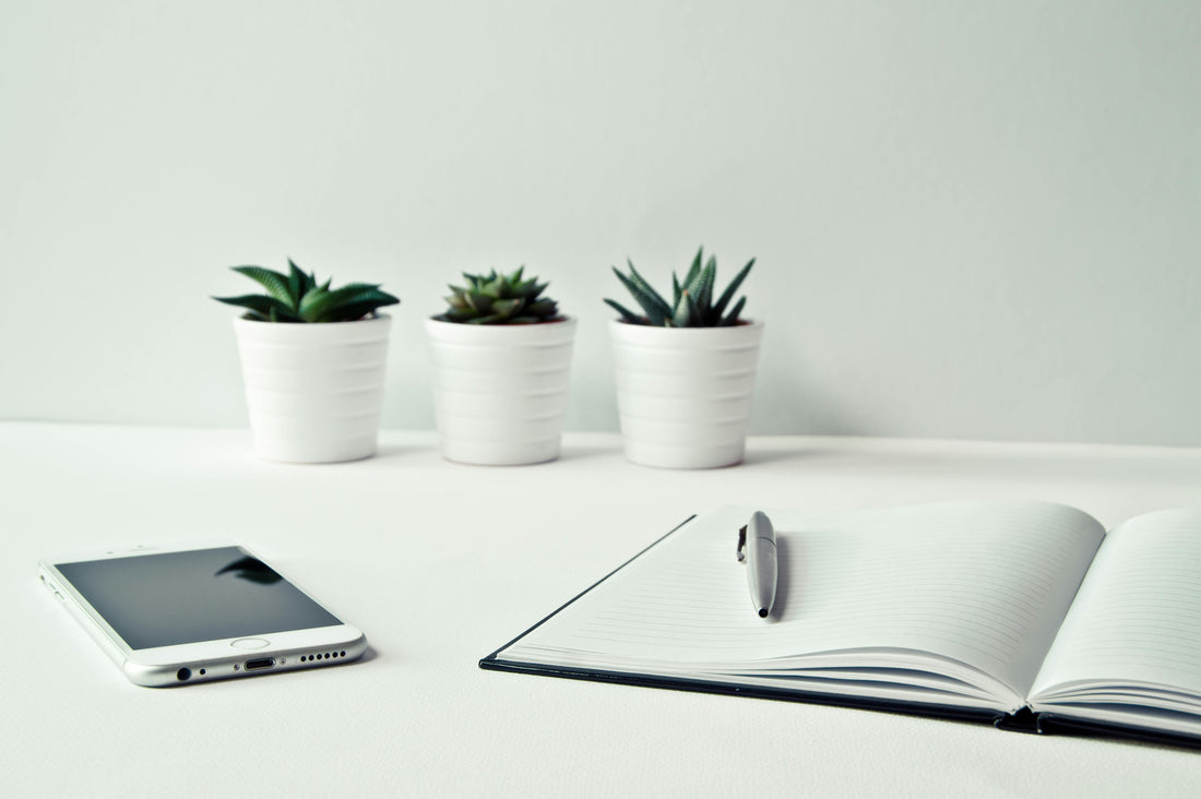 A white desk with a note book open at a blank page, with a pen sat on it. There is also a mobile phone and three small plants on the desk.